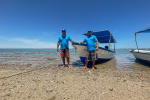 Two men stand on a beach in front of a fishing boat